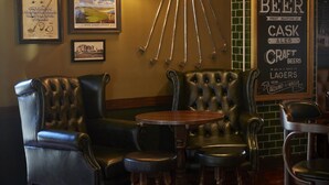 A bar area with leather chairs, a round wooden table, and a wall with framed pictures and a sign about beer. - Greyfriars Inn by Greene King Inns (St. Andrews)