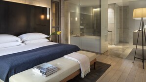 A modern hotel room with a large bed, a bedside table with books, a glass-enclosed shower area, and a standing lamp.