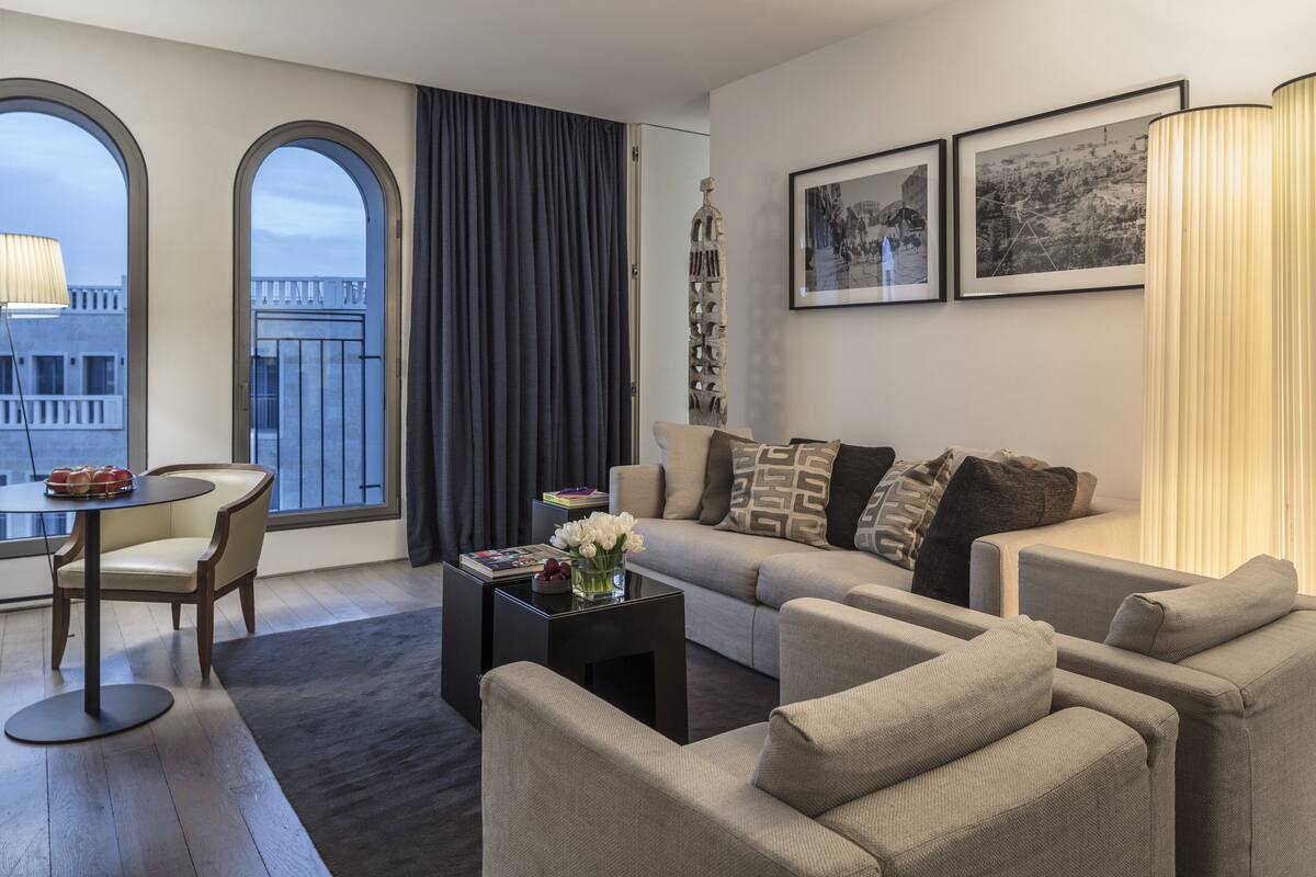 Un salon moderne comprenant un canapé, des fauteuils, une table basse et une table à manger. De grandes fenêtres sont ornées de rideaux, une sculpture décorative est présente, et des tableaux encadrés décorent le mur.