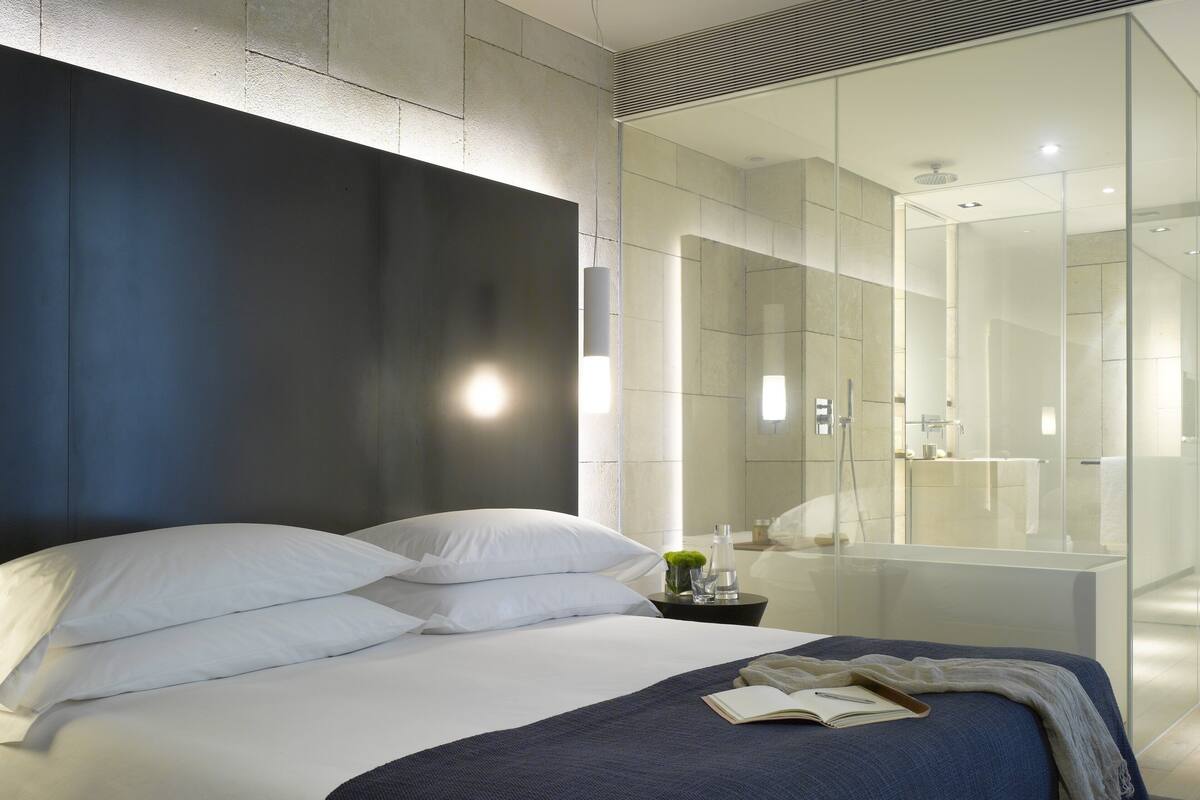 A modern hotel room with a large bed, a bedside table with a lamp, and a glass shower enclosure.