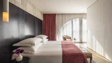 Une chambre d’hôtel moderne avec un grand lit, une table de chevet ornée d’un vase, une chaise et une vue sur l’extérieur à travers de fines rideaux.