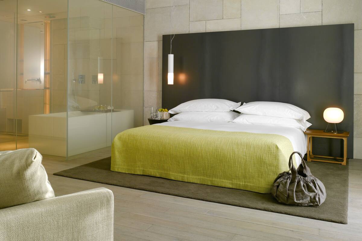 Une chambre d’hôtel moderne avec un grand lit, une table de chevet avec une lampe, une cabine de douche en verre et vue sur la salle de bain.