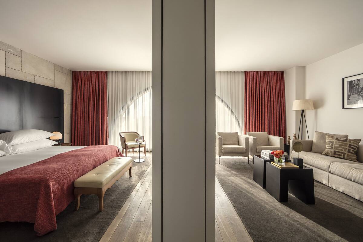 Une chambre d’hôtel avec un lit, une banquette, un canapé et une chaise. Il y a une fenêtre avec des rideaux et une lampe sur une table d’appoint.
