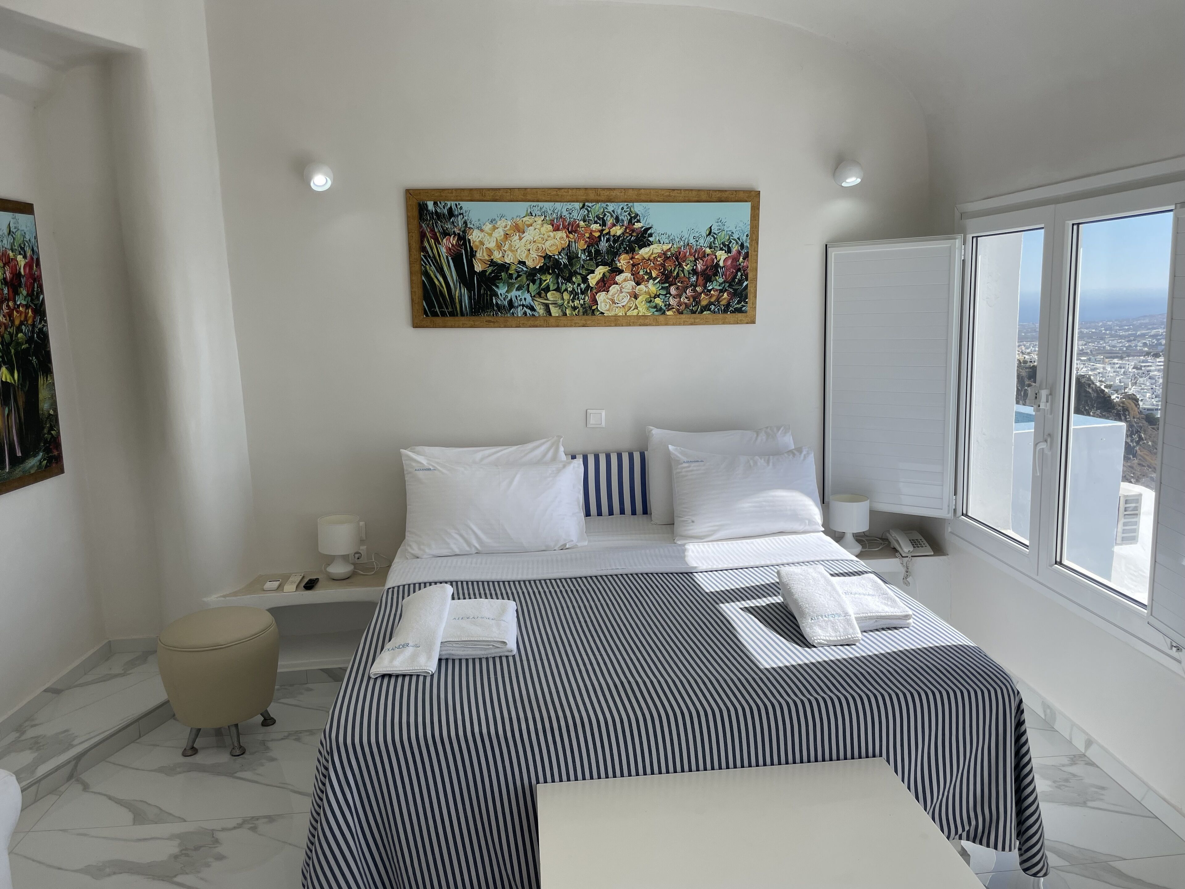 A hotel room with a bed, a painting, a window with a city view, and a small table with a lamp.