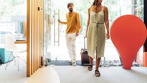 Un moderno vestíbulo de hotel con un separador de madera, una gran escultura abstracta roja y una puerta de vidrio que da al exterior.