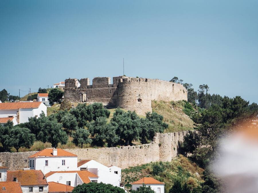 Um castelo de pedra histórico no alto de uma colina, rodeado por edifícios brancos e telhados de telha vermelha.