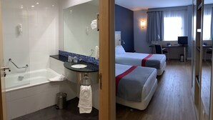 Habitación de hotel con bañera, lavamanos, espejo, dos camas, escritorio con computadora y ventana con cortinas.