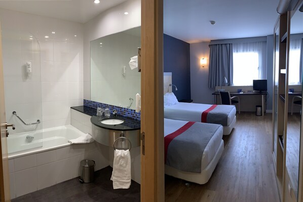 Habitación de hotel con bañera, lavamanos, espejo, dos camas, escritorio con computadora y ventana con cortinas.