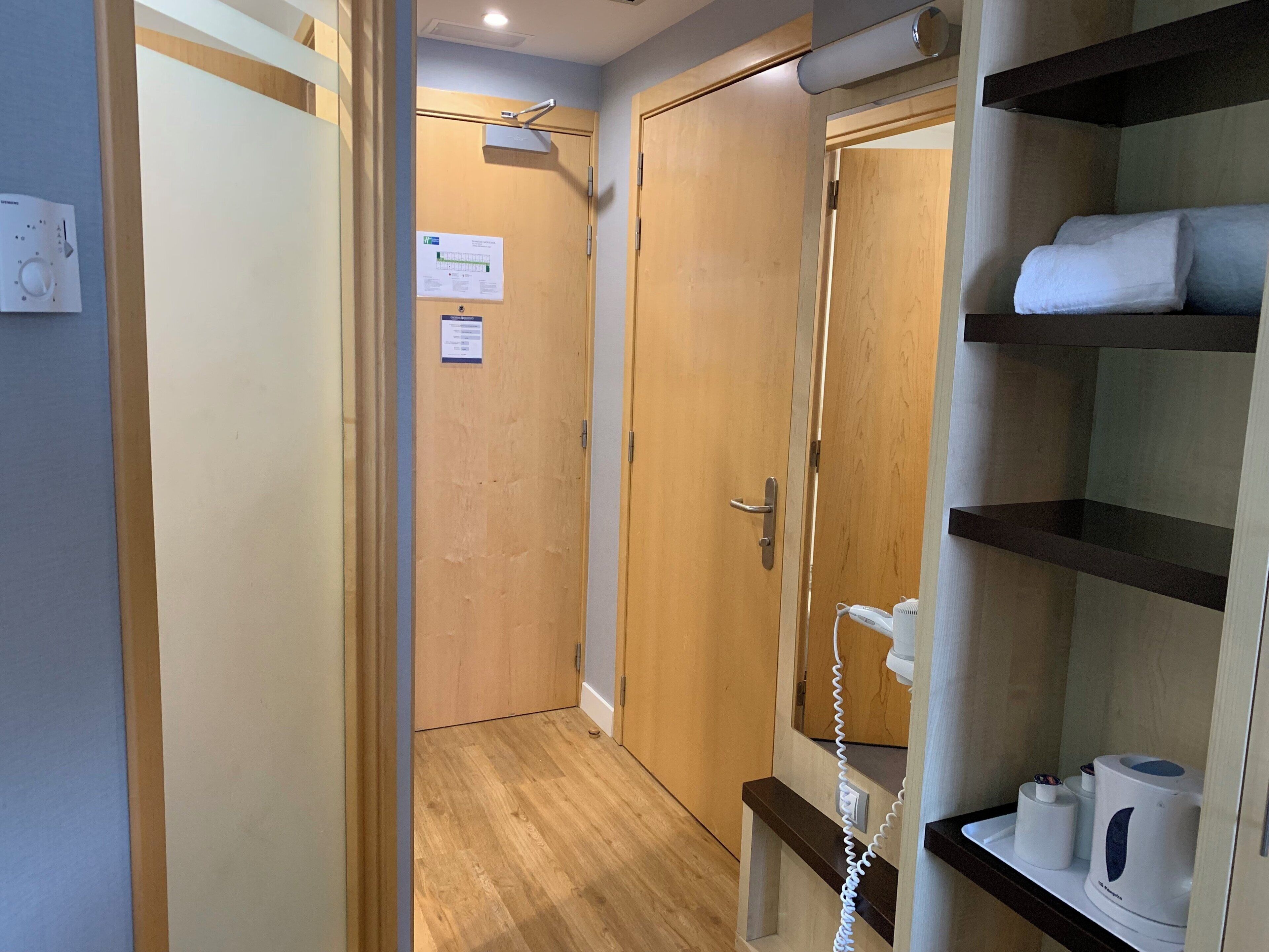Habitación de hotel con puerta de madera, pared blanca, suelo de madera, repisa con artículos de aseo y teléfono fijo en la pared.