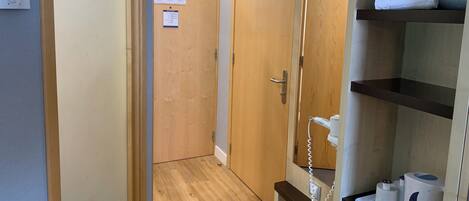 Habitación de hotel con puerta de madera, pared blanca, suelo de madera, repisa con artículos de aseo y teléfono fijo en la pared.