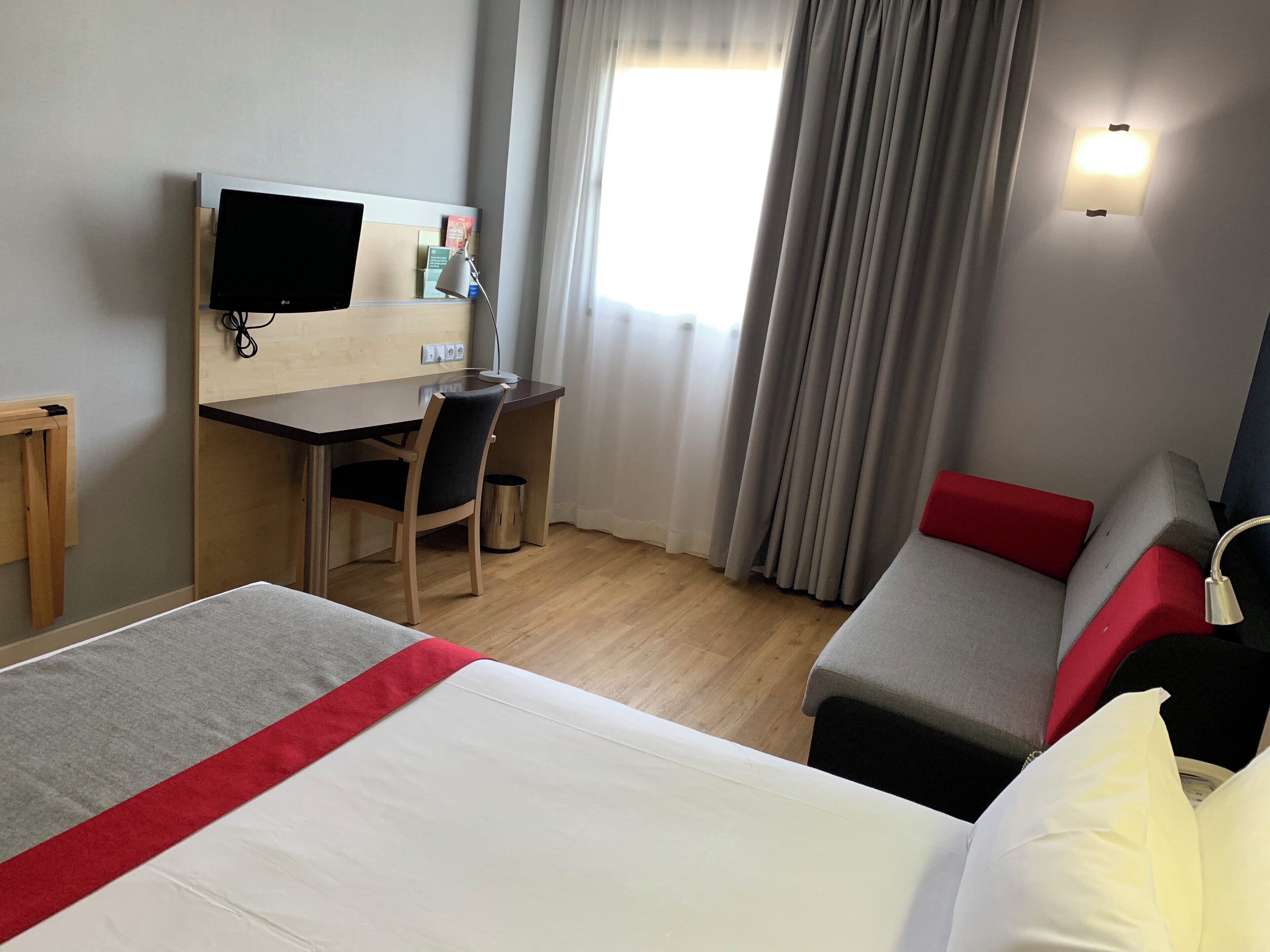 A hotel room with a bed, a desk, a chair, a TV, and a window with curtains.