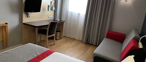 A hotel room with a bed, a desk, a chair, a TV, and a window with curtains.