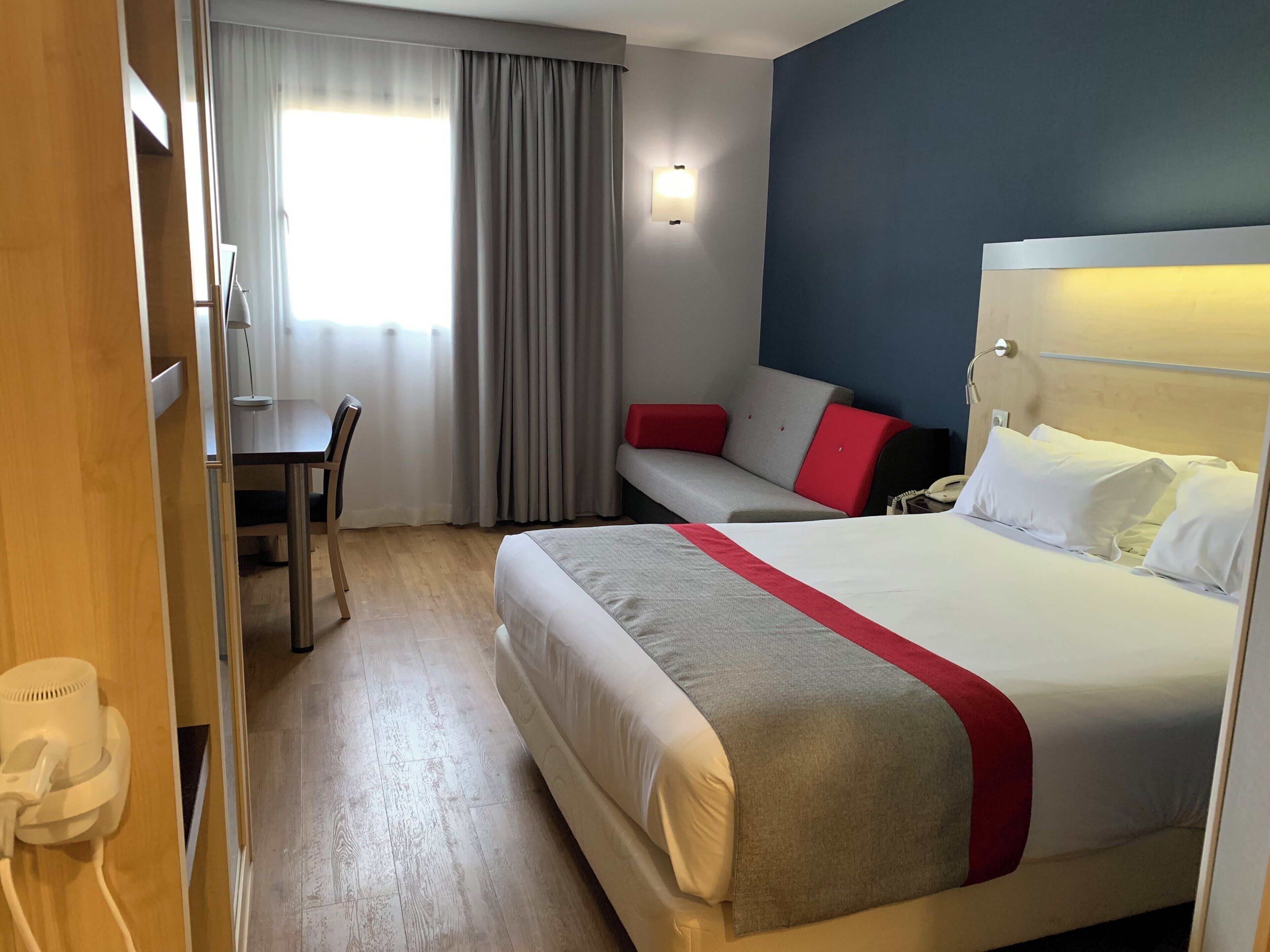A hotel room with a bed, a desk, a chair, a sofa, and a window with curtains.