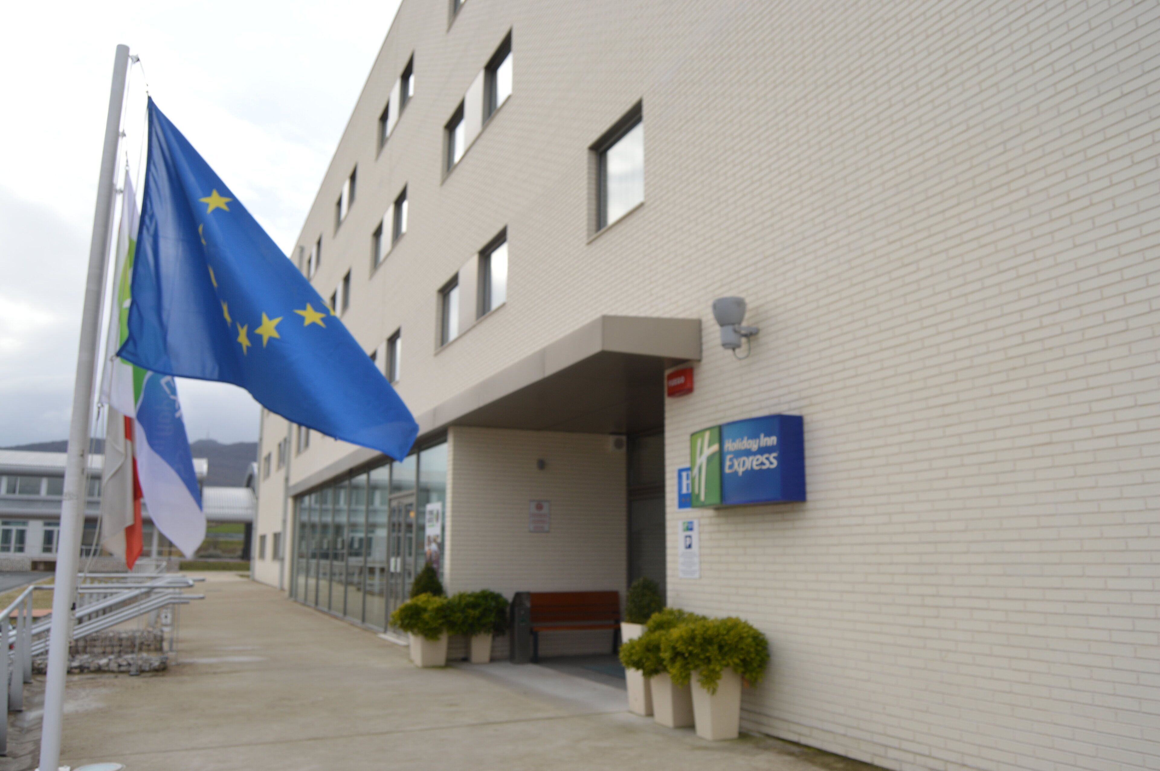 Een gebouw met een Holiday Inn Express bord, een vlaggenmast met verschillende vlaggen en plantenbakken bij de ingang.