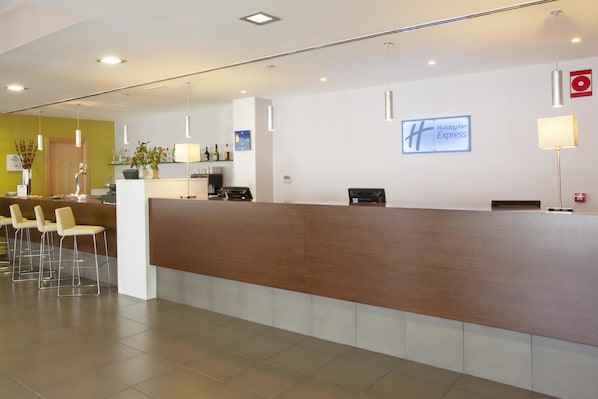 Una zona de recepción con un mostrador de madera, varios taburetes tipo bar, una planta en maceta y un cartel de Holiday Inn Express en la pared.