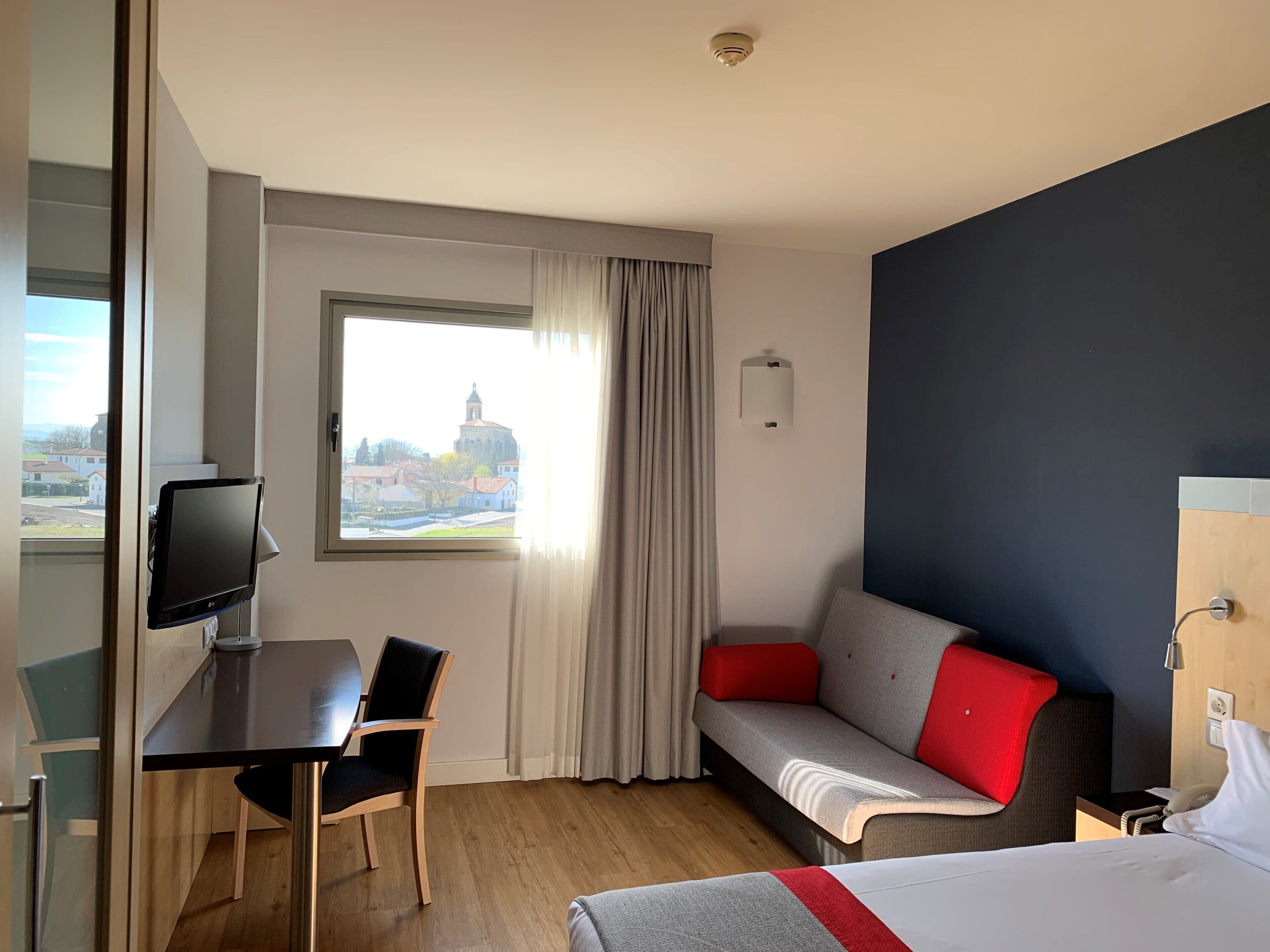 A hotel room with a bed, a desk with a chair, a sofa with red cushions, a television, and a window with curtains.
