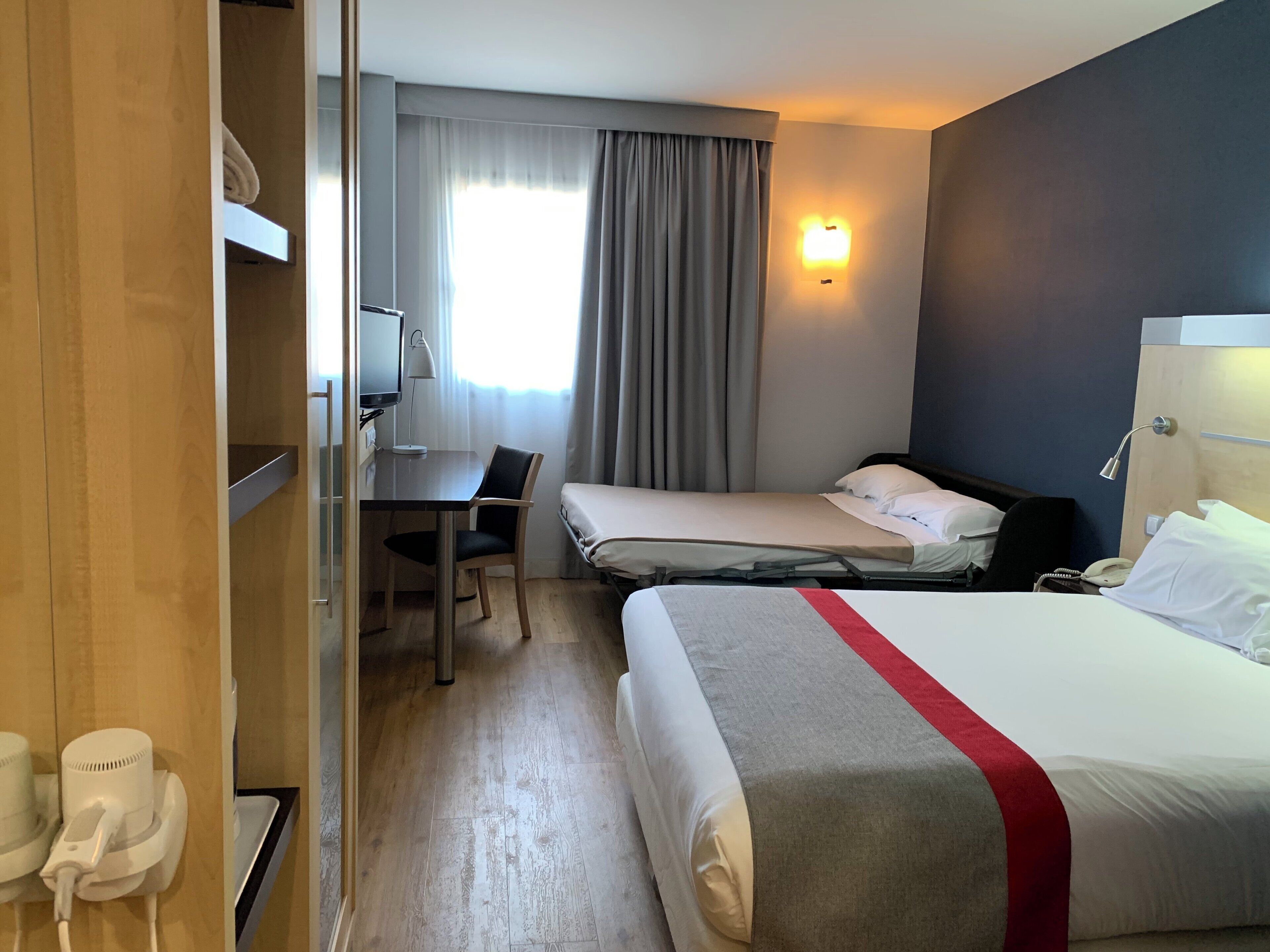A hotel room with a bed, a desk, a chair, a telephone, and a window with curtains.