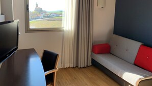 Habitación con televisor, escritorio de madera, una silla, sofá con cojines rojos, ventana con cortinas y vista a edificios.
