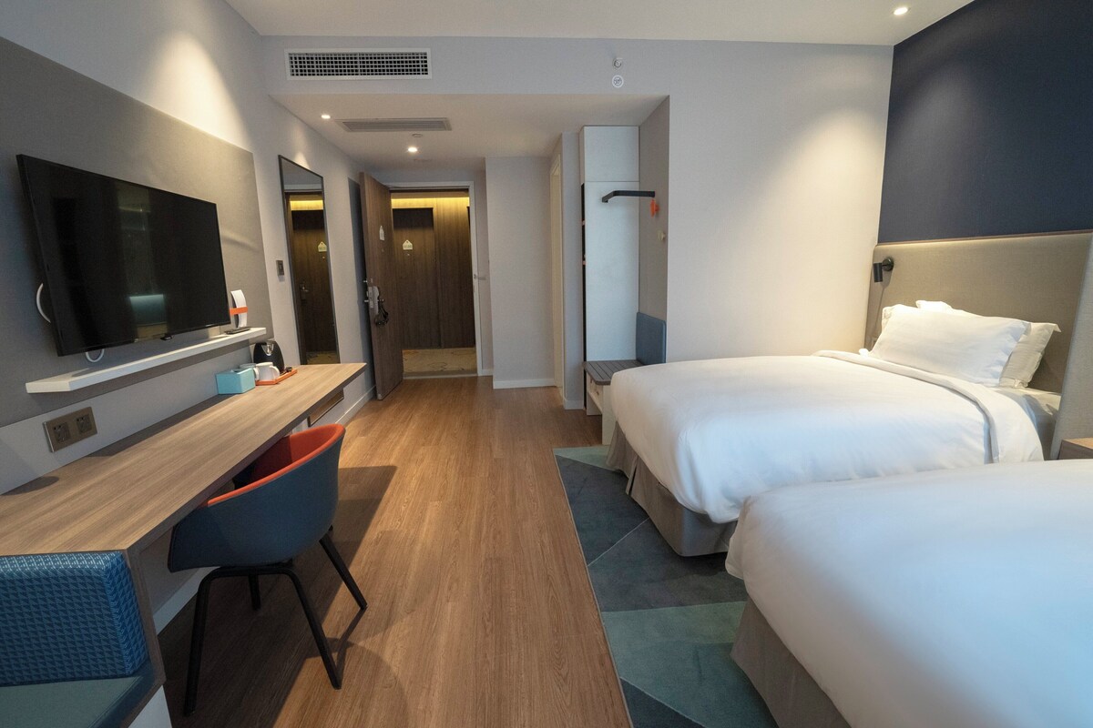 A hotel room with two beds, a desk, a chair, a TV, and a bathroom door.
