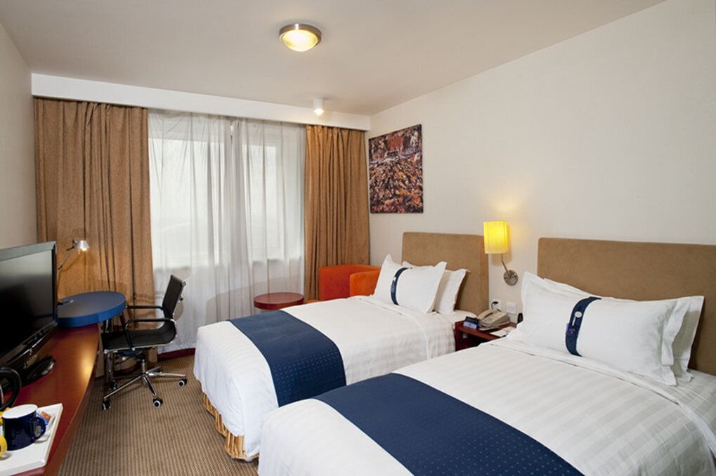 A hotel room with two beds, a desk with a computer, a chair, a TV, and a lamp.