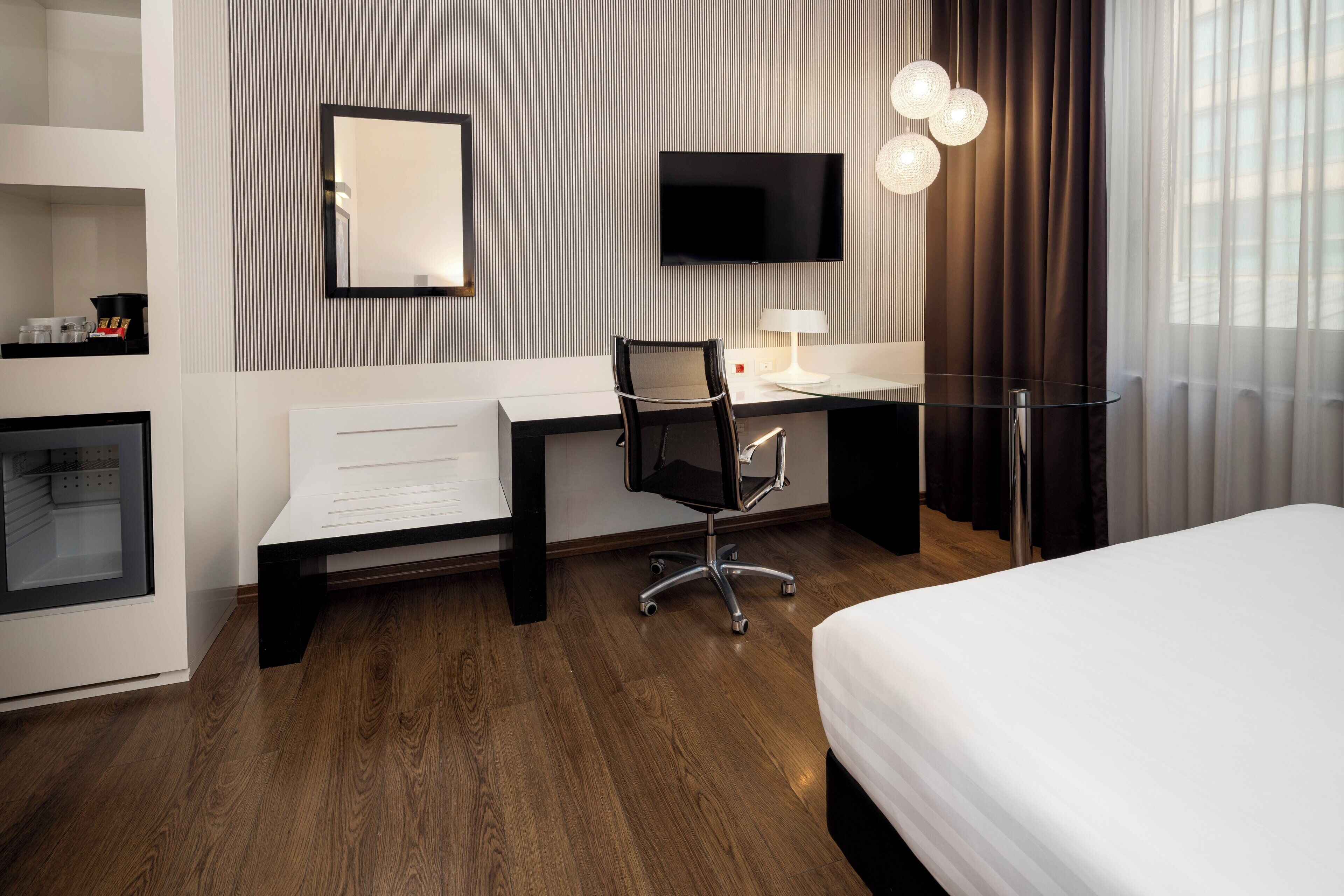 A modern hotel room with a wooden floor, a desk with a chair, a bed, a TV mounted on the wall, and a bedside lamp.