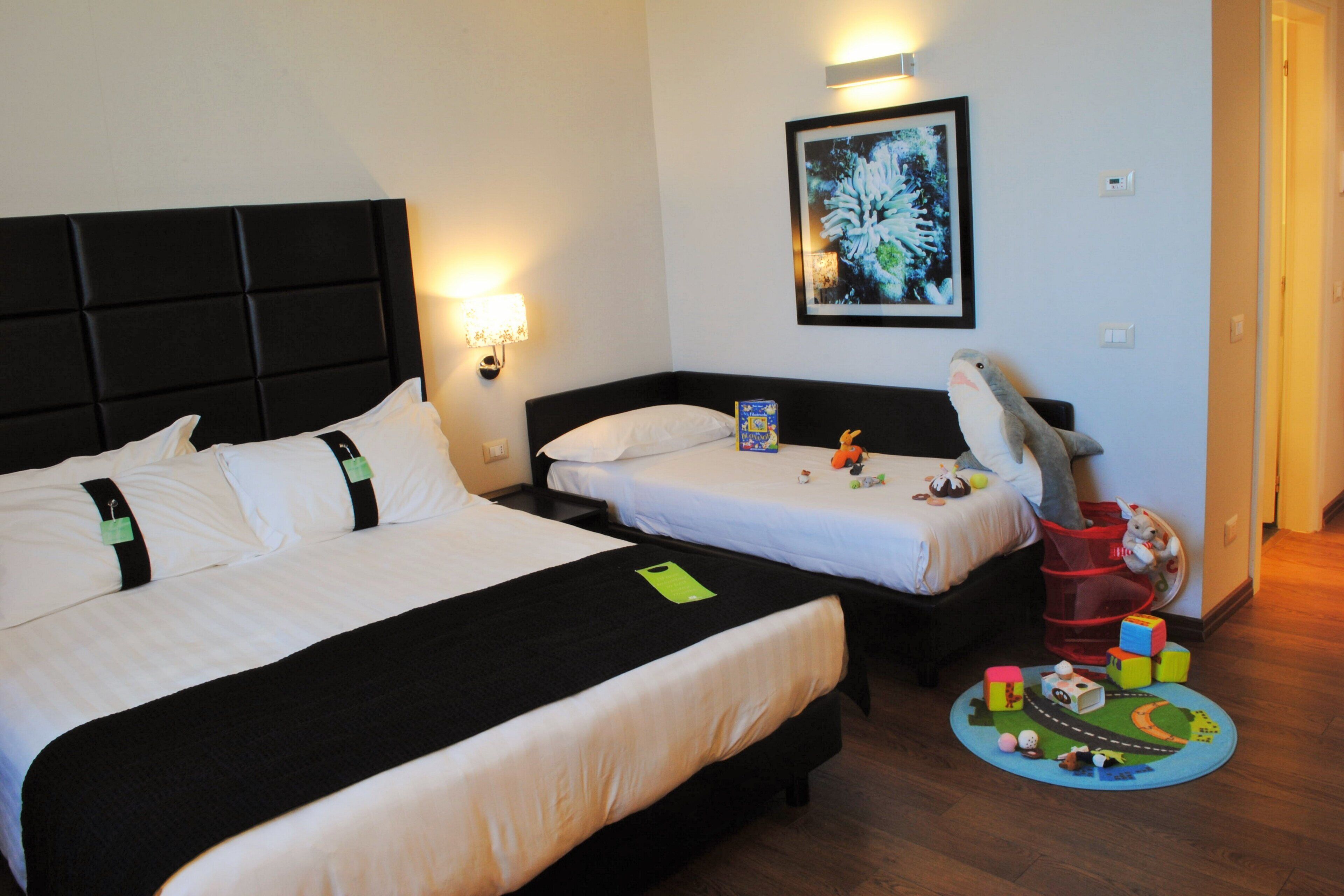 A hotel room with two beds, a nightstand, a framed picture on the wall, and a children's play area with toys.