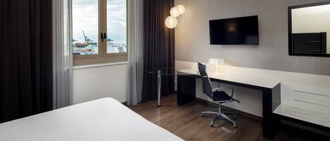 A hotel room with a large bed, a desk with a chair, a TV mounted on the wall, and a window with curtains.