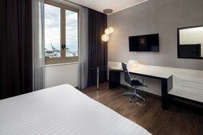 A hotel room with a large bed, a desk with a chair, a TV mounted on the wall, and a window with curtains.