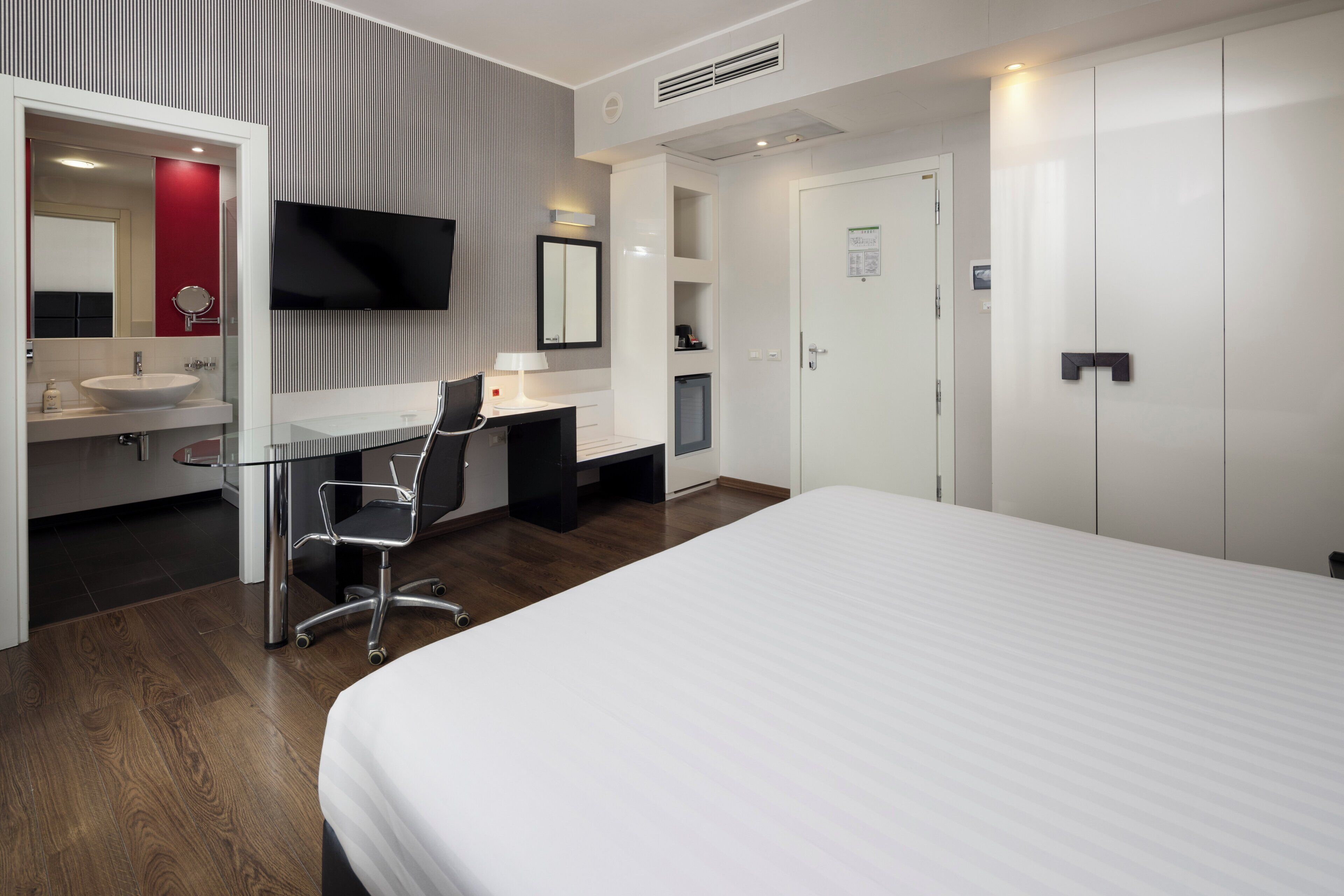 A hotel room with a bed, a desk with a chair, a TV, a mirror, a sink, and a wardrobe.