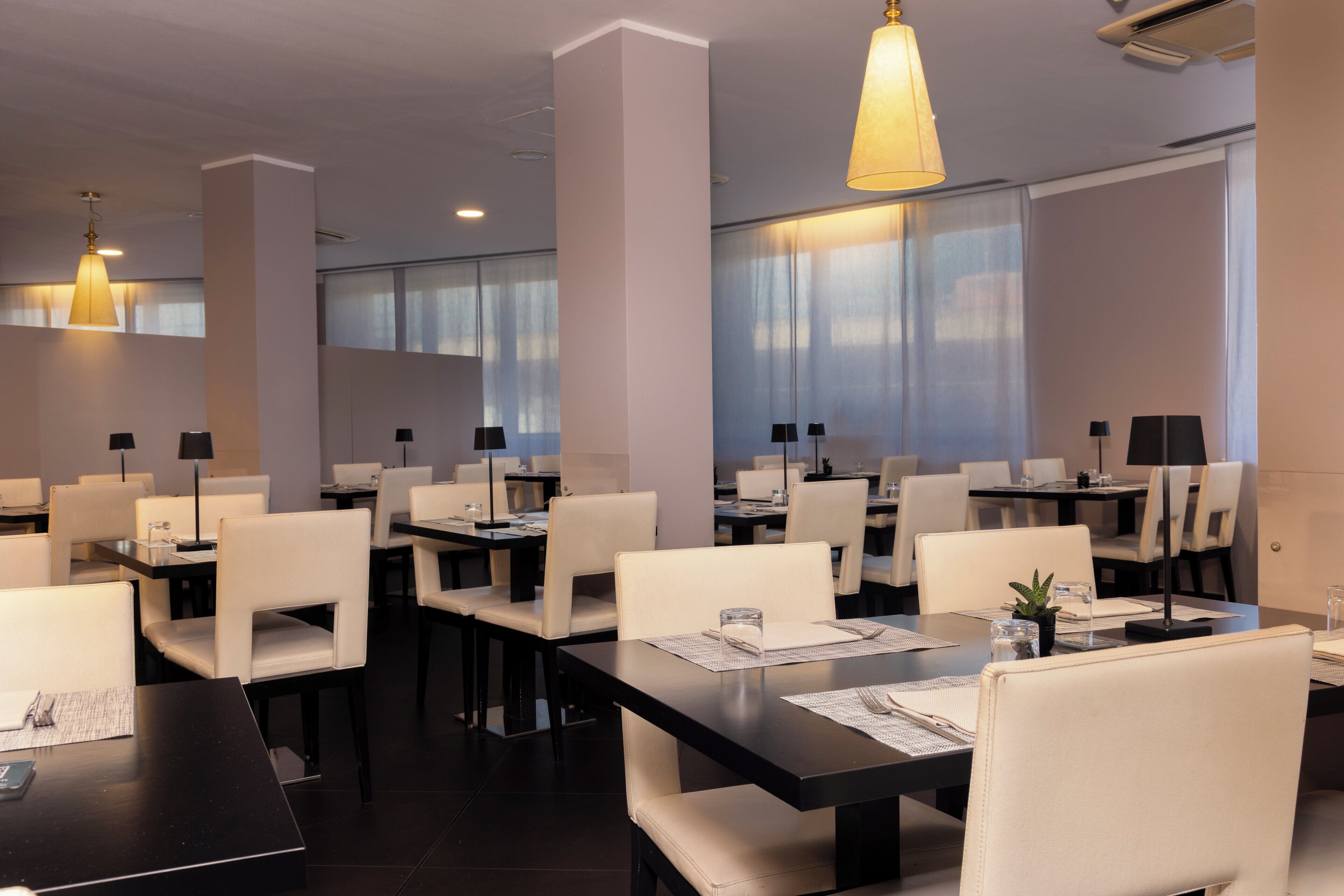 Un comedor moderno con mesas y sillas, ventanas amplias e iluminación colgante.