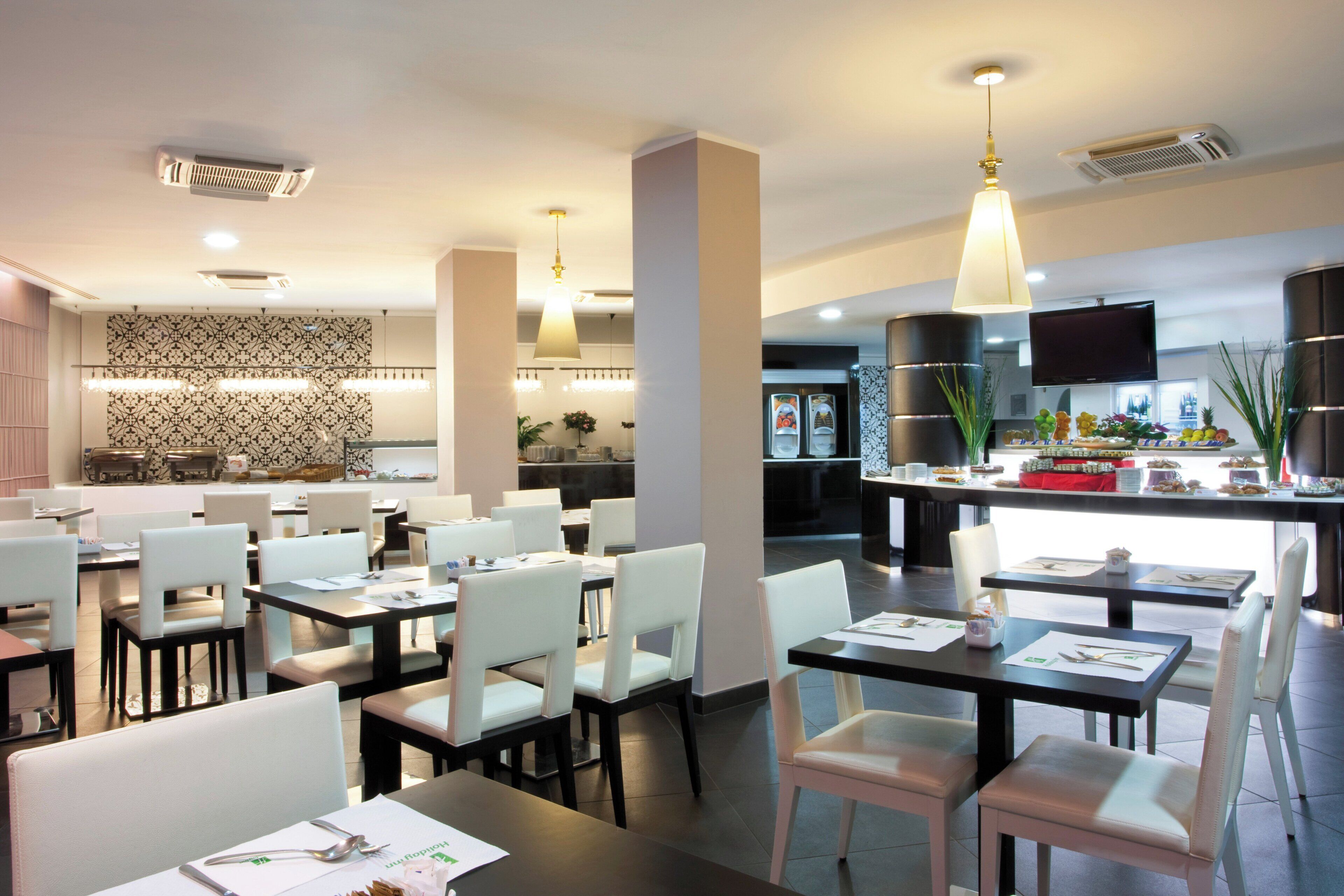 A modern dining area with tables and chairs, a kitchen in the background, and a buffet counter with food items.