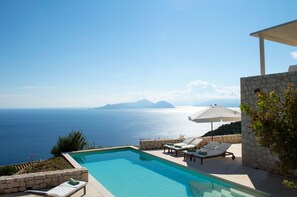 A pool area with lounge chairs and umbrellas overlooking a body of water with distant landforms. - Urania Luxury Villas (Lefkada)
