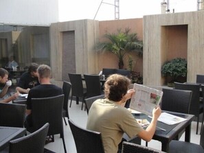 An outdoor café with wicker chairs and tables, a palm plant, and a concrete wall. - Hotel Chanchal Deluxe (New Delhi)