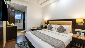 A hotel room with a large bed, a desk, a chair, a small table with a fruit bowl, a nightstand with a lamp, and a ceiling fan. - Hotel Chanchal Continental (New Delhi)