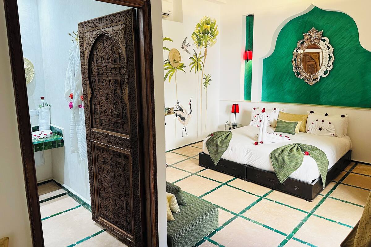 A hotel room with a large ornate wooden door, a green accent wall, a bed with green and white bedding, and a tiled floor.