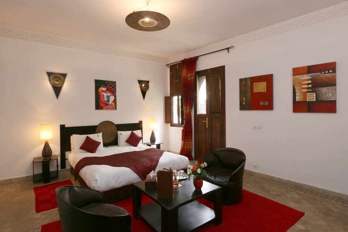 A hotel room with a bed, a desk, two chairs, a red rug, and artwork on the walls.