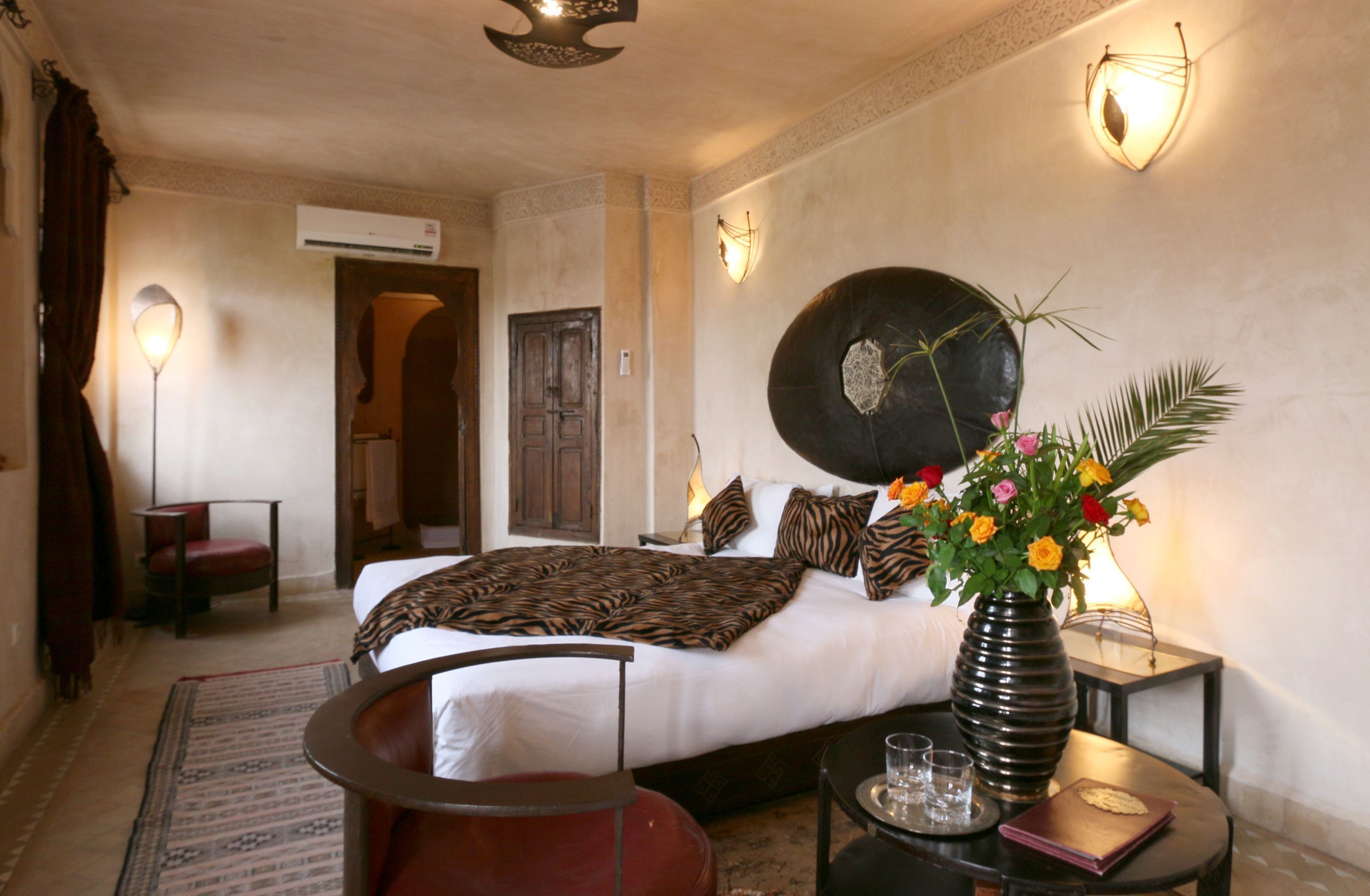 Una habitación de hotel con una cama, una zona de estar con una silla, una mesa con un jarrón de flores y una lámpara fijada a la pared.