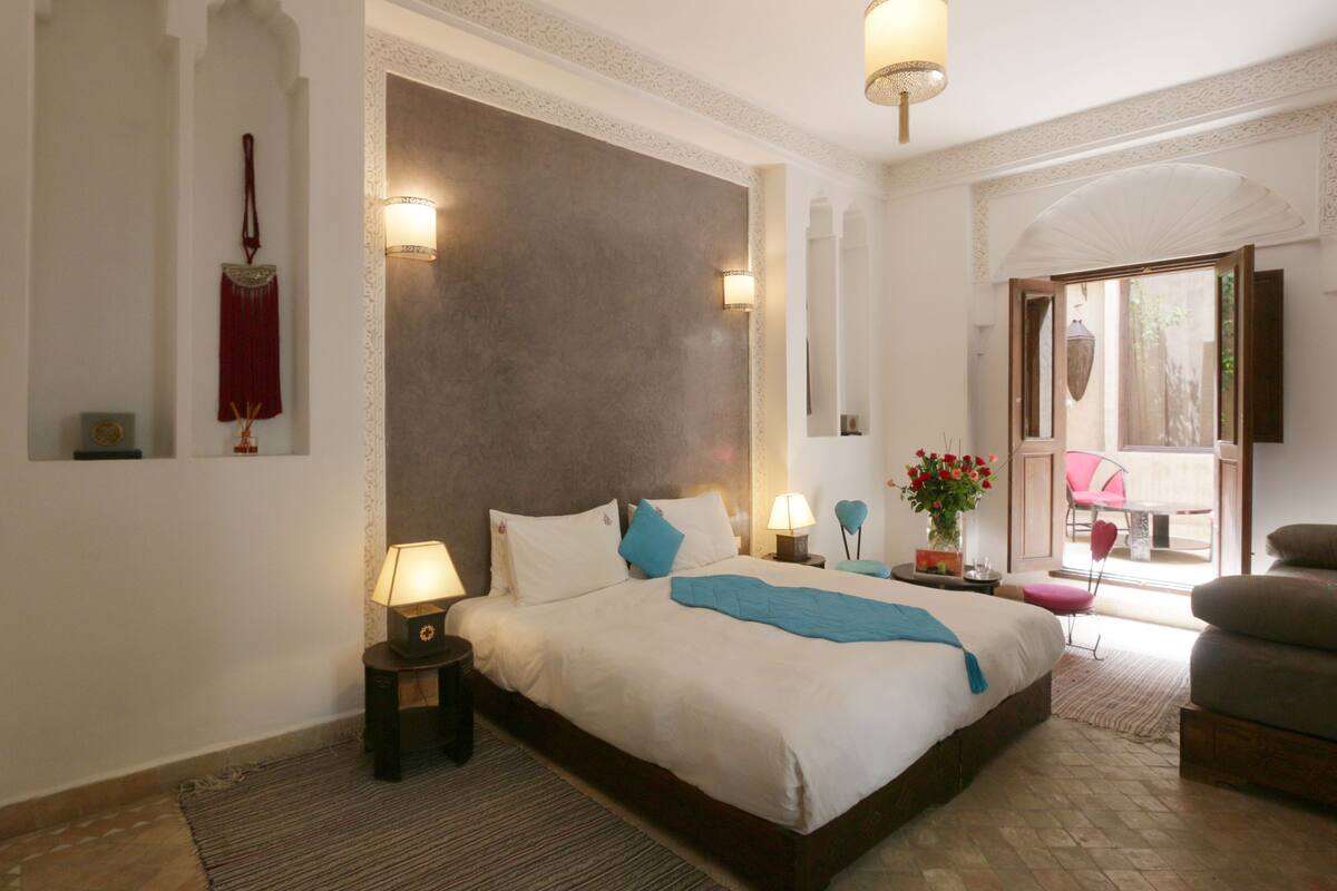 A bedroom with a large bed, two bedside tables with lamps, a red decorative item on the wall, and a view of the outside through a glass door.