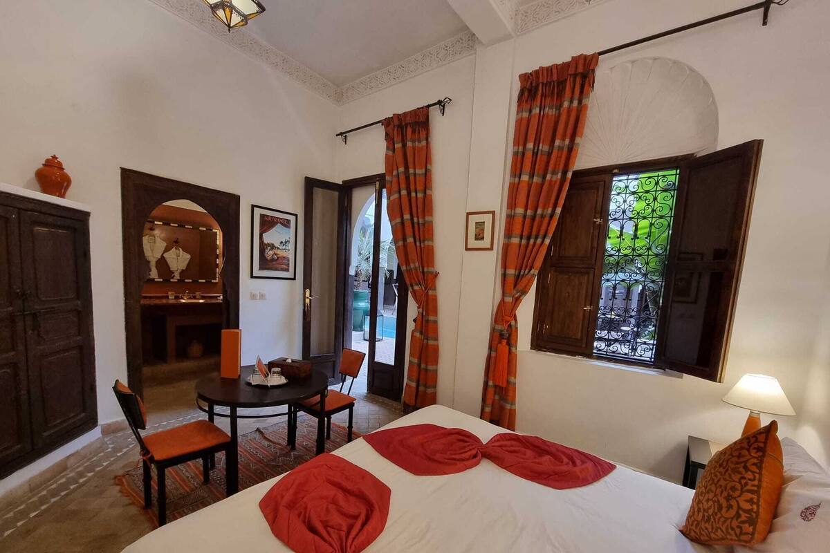 A hotel room with a bed, a dining table with chairs, a small table with a vase, a lamp, and a window with curtains.