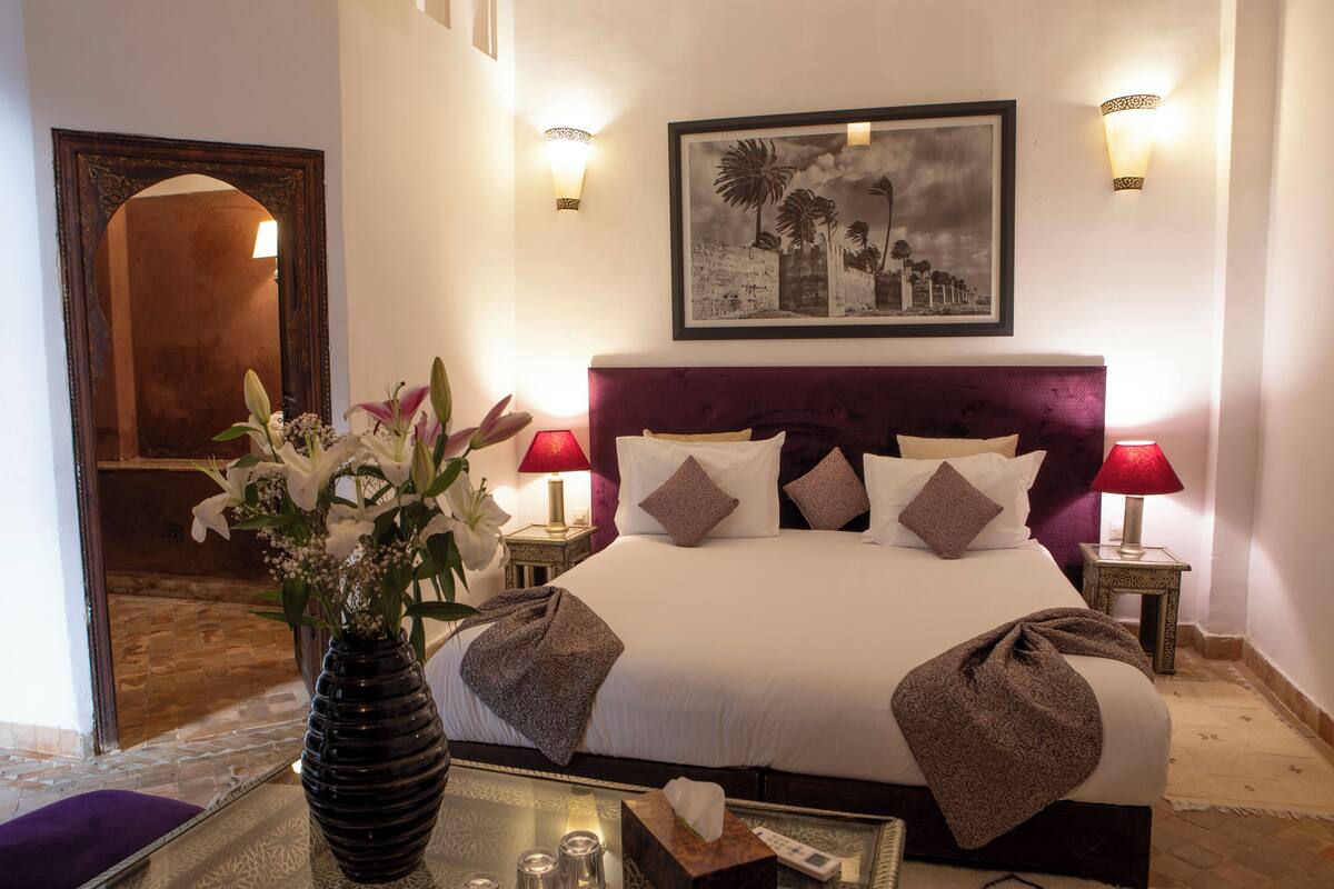 A hotel room with a large bed, two bedside tables with lamps, a vase with flowers, a framed picture on the wall, and a mirror in the corner.