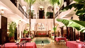 A hotel lobby with a central pool, multiple dining tables with red tablecloths, and a staircase leading to an upper level.
