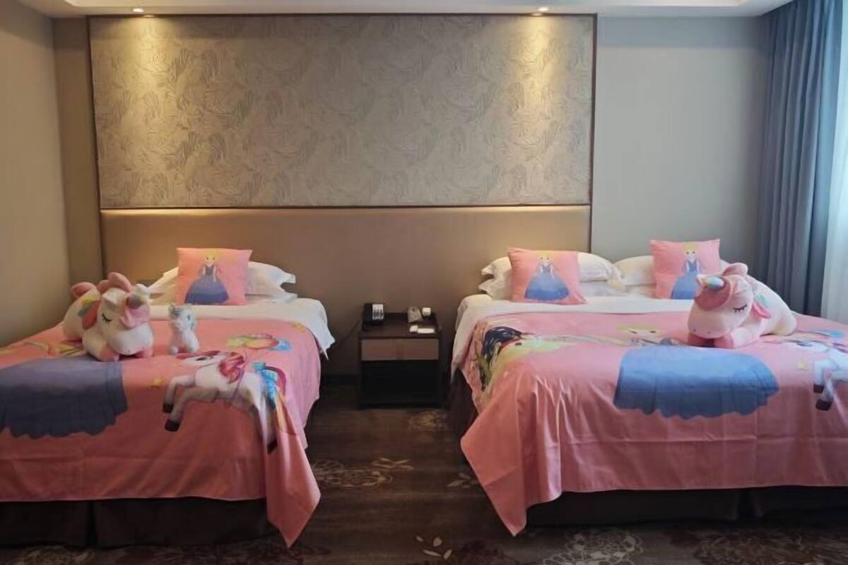 A hotel room with two beds, each with a pink bedspread and a unicorn plush toy. There is a nightstand between the beds with a lamp and a phone.