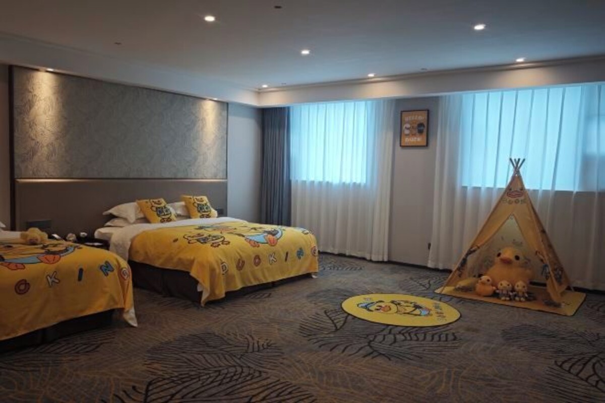 Quarto de hotel com duas camas, uma janela grande com cortinas, cabeceira fixada na parede, decoração tipi e tapete com padrão de folhas.