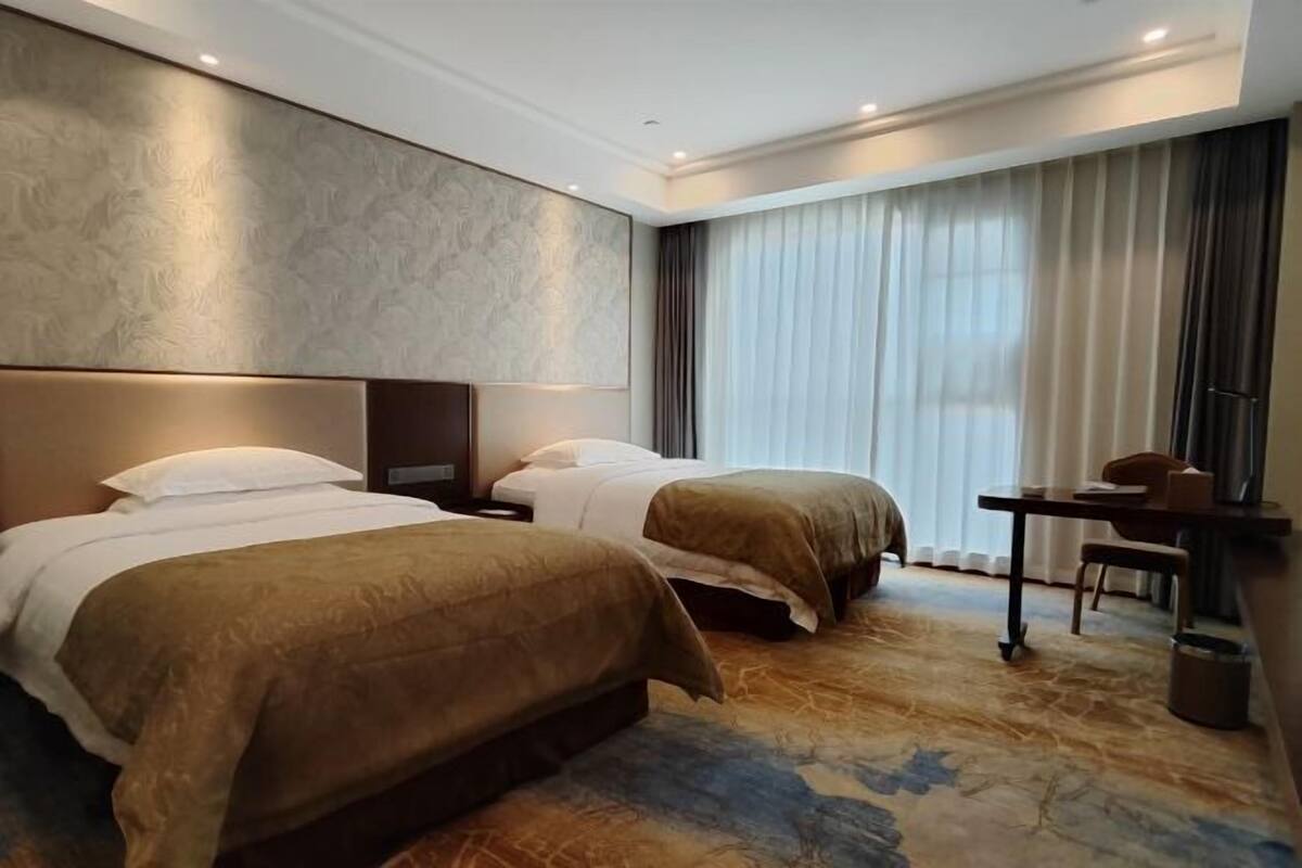 A hotel room with two beds, a desk, and a large window with curtains.