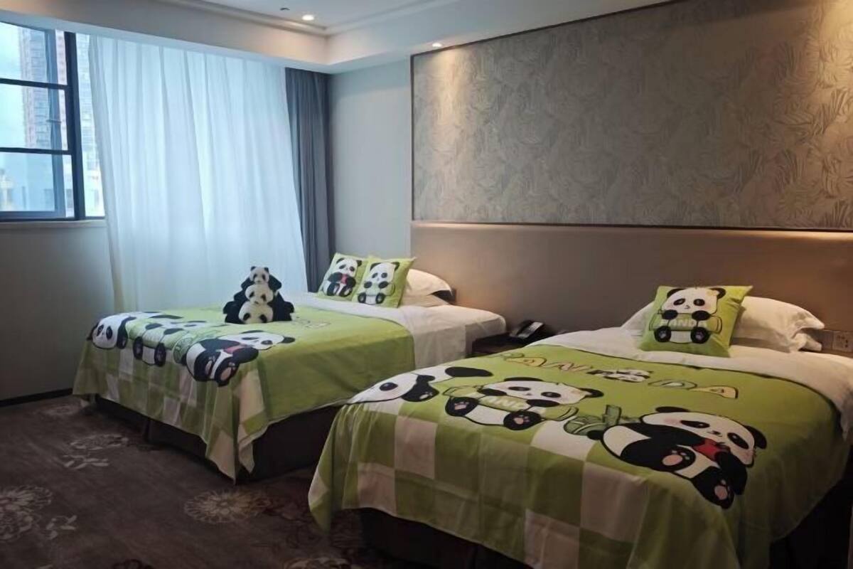 A hotel room with two beds, each with a green and white checkered bedspread featuring panda designs, a large window with sheer curtains, and a textured wall panel above the beds.