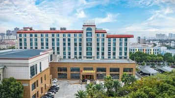 A multi-story hotel with a parking lot, surrounded by greenery and a cityscape in the background.