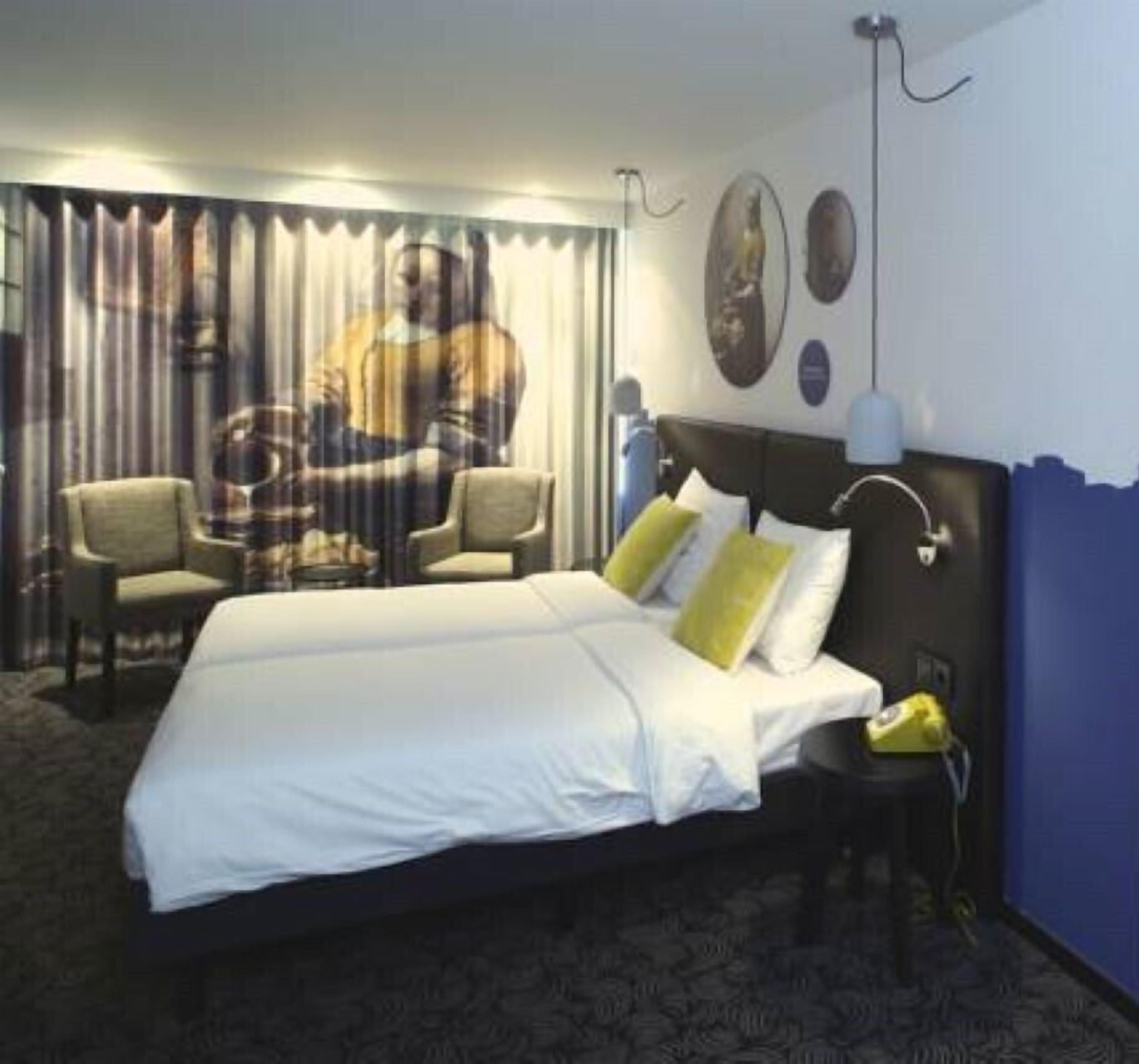 Une chambre d’hôtel avec un grand lit, deux chaises, un bureau et une lampe fixée au mur.