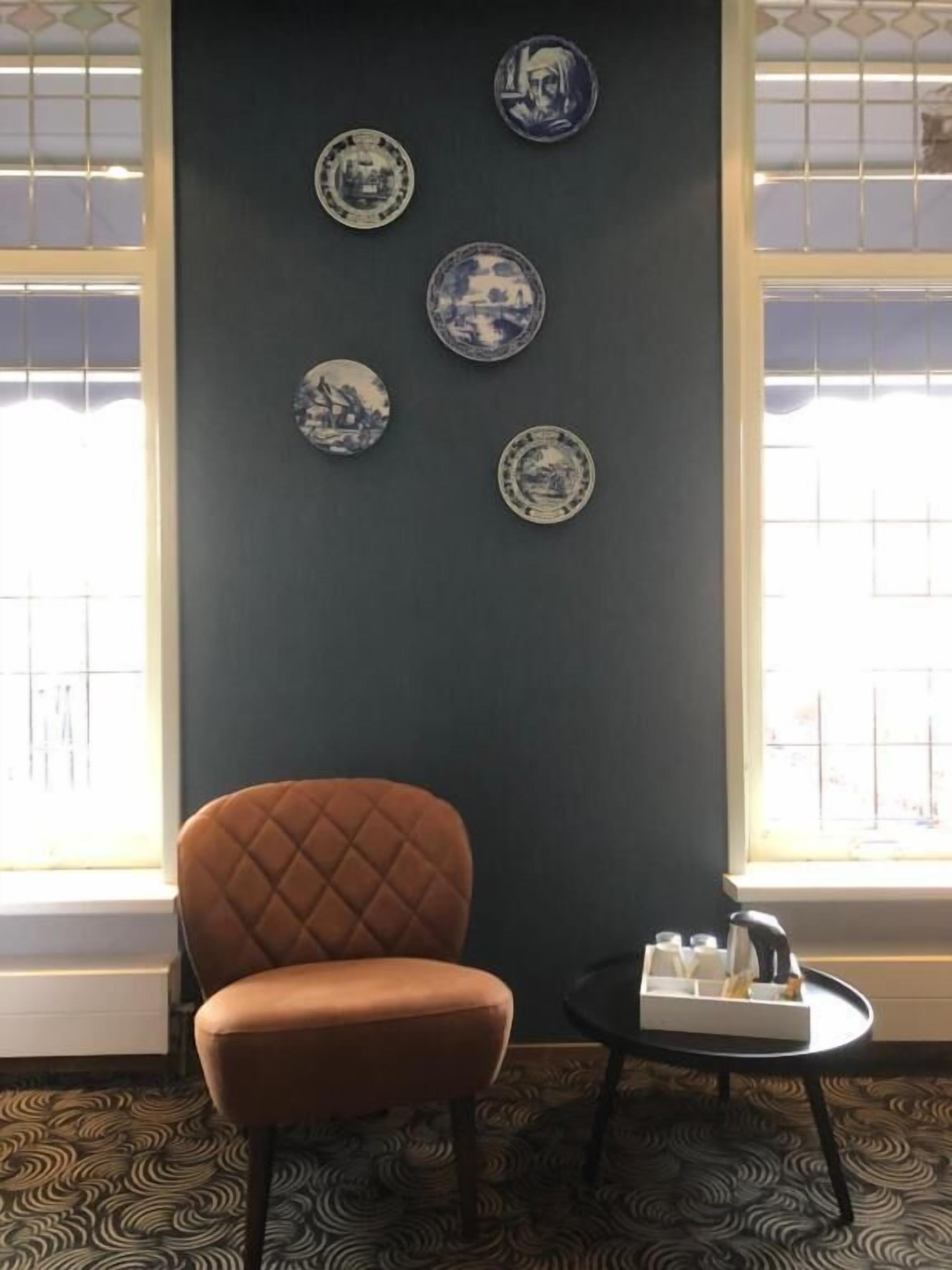 Een kamer met een donkere muur, een bruine stoel, een klein rond tafeltje met een witte dienblad, en vier decoratieve borden aan de muur.