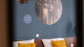 Quarto de hotel com duas camas, cada uma com colchão branco e travesseiro amarelo. Duas decorações circulares na parede, uma retratando uma cena histórica e outra um grupo de pessoas.