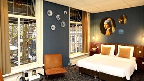 A hotel room with a large bed, a chair, a small table, and a window with a view of buildings. - Hotel de Koophandel (Delft)